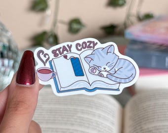 May include: A white sticker with a blue book, a gray cat sleeping on top of it, and a red cup of coffee. The sticker says "Stay Cozy" in pink letters.