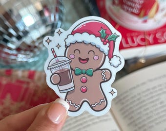 May include: A sticker featuring a smiling gingerbread man wearing a Santa hat and holding a drink. The gingerbread man has a green bow tie and red buttons. The drink is in a clear cup with a straw.