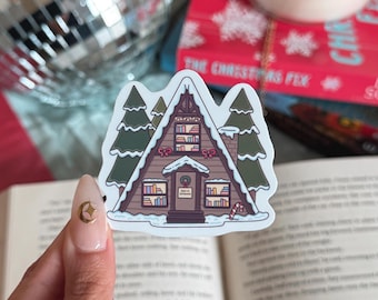 May include: A sticker featuring a brown A-frame cabin with snow-covered roof and evergreen trees. The cabin has windows with books, a door with a wreath, and a candy cane. The text "Open for Christmas" is on the door.