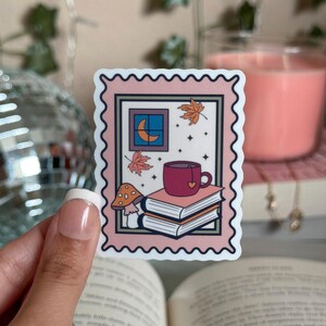 May include: A pink and white sticker with a vintage postage stamp design. The sticker features a cup of tea, a stack of books, a mushroom, and a window with a crescent moon.