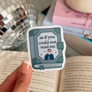 May include: A sticker featuring a book design with the text "as if you could out read me." The book is light blue with darker blue accents and a bookmark. A cartoon figure wearing sunglasses is inside the book, with small hearts around it.