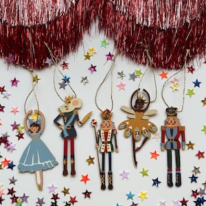 May include: Five flat wooden hanging ornaments featuring characters from The Nutcracker: a girl in a blue dress, a mouse king, a nutcracker soldier with a staff, a fairy, and a soldier, all suspended by gold strings amidst colourful star confetti.