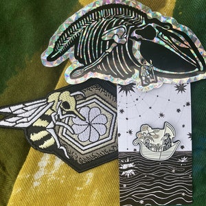 May include: Three embroidered patches featuring a bee, a flower, and a whale skeleton. The patches are on a green and yellow tie-dye fabric background.