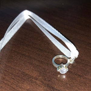 May include: A silver ring with a large clear stone, attached to a white ribbon.