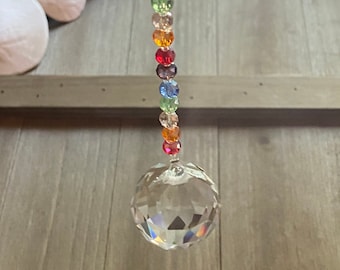 May include: A hanging suncatcher featuring a clear, multifaceted crystal sphere suspended from a vertical strand of small, colorful beads. The beads are arranged in a repeating sequence of purple, blue, green, clear, orange, and red hues.