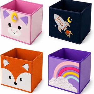 Puede incluir: Un conjunto de cuatro cajas de almacenamiento cuadradas de tela. Una es rosa con cara de unicornio, una azul oscuro con un cohete, una naranja con cara de zorro y una morada con un diseño de arcoíris y nube. Cada caja tiene la parte superior abierta.