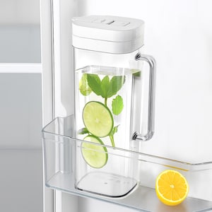May include: A clear rectangular water pitcher with a white lid and handle, filled with water, lime slices, and mint leaves. The pitcher is in a refrigerator shelf next to a half-cut yellow lemon.