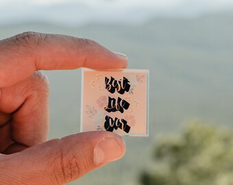 May include: A small, square sticker with the words "Best Day Ever" in black script. The sticker has a peach background with floral accents. The sticker is held between two fingers against a blurred outdoor background.