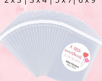 May include: A set of clear favor bags with pink and red labels that say "A little sweetheart is on the way!" with the name "Baby Marlene" and the date "May 2024". The bags are available in four sizes: 2x3, 3x4, 5x7, and 6x9.