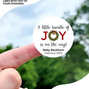 May include: A white circle sticker with a red and green wreath with gold bells and the words "A little bundle of JOY is on the way!"  The sticker also includes the text "Baby Beckham February 2024". The sticker is available in sizes 1 inch, 1.5 inches, 1.75 inches, 2 inches, 2.25 inches, and 3.33 inches.