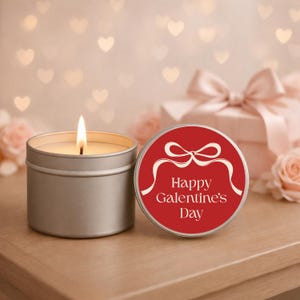 May include: A lit silver candle next to a red tin with the text "Happy Galentine's Day" and a white bow design. A gift box with a pink bow and pink roses are in the background. The candle is on a wooden surface.