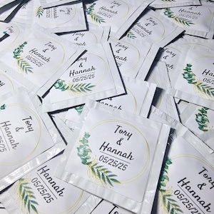 May include: A pile of white favor bags with a green and gold floral design and the text "Tory & Hannah 05/25/25" printed on each bag.