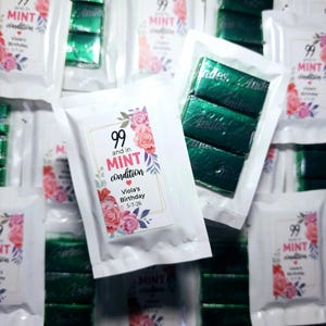 May include: A white bag with a pink and green floral design sticker that says "99 and in MINT condition" with the text "Viola's Birthday 5-7-26". The bag contains three individually wrapped green chocolate bars.