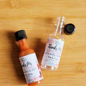 May include: Two small glass bottles of hot sauce with white labels. The labels have pink floral designs and the text "Thanks for Spicing Up Our Special Day!" with the date "9.29.2022" and the names "Bob & Mary".