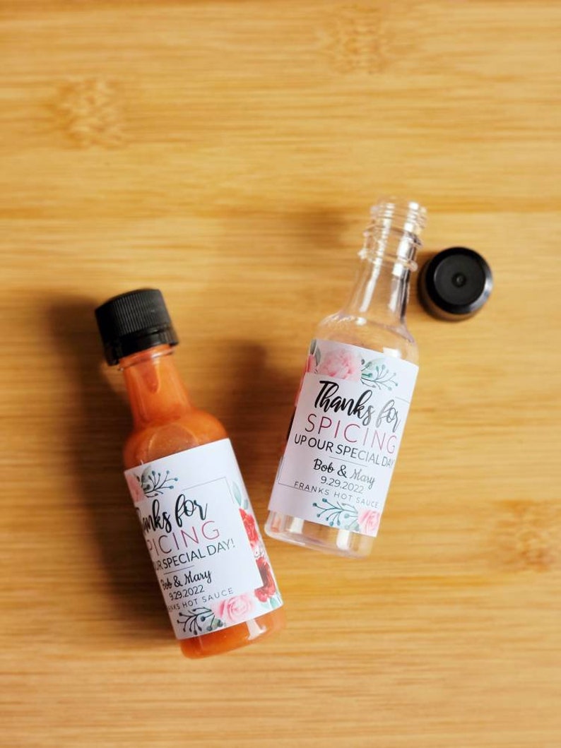 May include: Two small glass bottles of hot sauce with white labels. The labels have pink floral designs and the text "Thanks for Spicing Up Our Special Day!" with the date "9.29.2022" and the names "Bob & Mary".