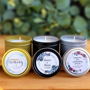 May include: Three small candle tins with personalized labels. The gold tin has a blue and gold floral design with the text "Thank you for coming Levi & Shaysea 02.26.2022". The black tin has a blue and gold floral design with the text "Laura & Kevin 2.6.2022". The silver tin has a red and gold floral design with the text "Thank you for celebrating with us! Ricardo & Rochelle 02.12.2022".