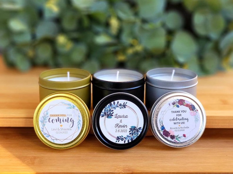 May include: Three small candle tins with personalized labels. The gold tin has a blue and gold floral design with the text "Thank you for coming Levi & Shaysea 02.26.2022". The black tin has a blue and gold floral design with the text "Laura & Kevin 2.6.2022". The silver tin has a red and gold floral design with the text "Thank you for celebrating with us! Ricardo & Rochelle 02.12.2022".