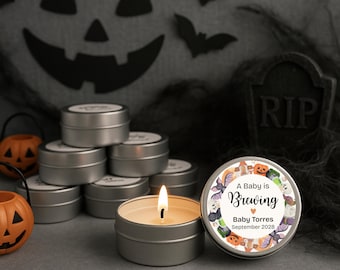 May include: A lit candle in a silver tin, with a label that reads "A Baby is Brewing" surrounded by Halloween-themed illustrations. Other silver tins are stacked in the background, along with decorative pumpkins and bats.
