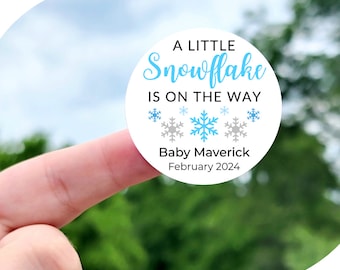 May include: A white circle sticker with blue and gray snowflakes and the text "A Little Snowflake Is On The Way" with the name "Baby Maverick" and the date "February 2024". The sticker is 1 inch, 1.5 inches, 1.75 inches, 2 inches, 2.25 inches, or 3.33 inches in diameter.