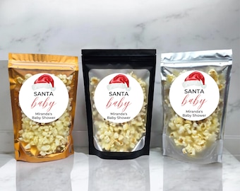 May include: Three clear plastic bags filled with popcorn, each with a round white sticker featuring a red Santa hat and the text "SANTA baby Miranda's Baby Shower". The bags are on a white marble surface.