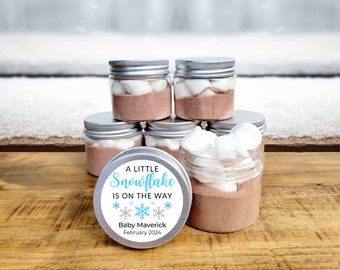 May include: A little snowflake is on the way baby shower favor jars with hot chocolate mix and marshmallows. The jars have a blue and silver snowflake design with the text "A little snowflake is on the way" and "Baby Maverick February 2024".