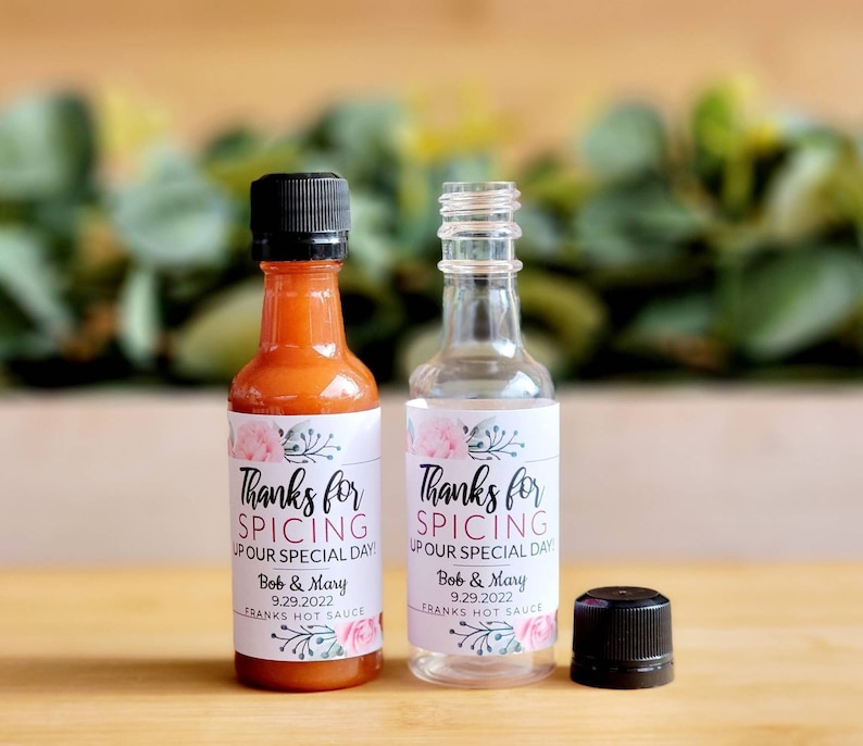 May include: Two small glass bottles with labels that say "Thanks for Spicing Up Our Special Day!" with a floral design. The labels also include the names "Bob & Mary" and the date "9.29.2022". The bottles are filled with hot sauce and Franks Hot Sauce.