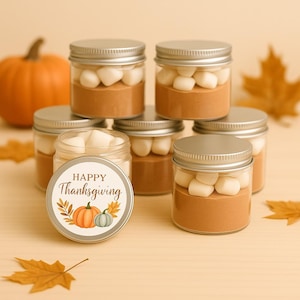 May include: A collection of small glass jars with silver lids, filled with a brown powder and white marshmallows. A circular label reads "Happy Thanksgiving" with pumpkin and leaf illustrations. An orange pumpkin and fall leaves are in the background.