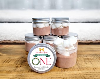 May include: A set of six clear glass jars with silver lids filled with hot chocolate mix and marshmallows. Each jar has a festive Christmas-themed label that says "Oh what fun it is to be one" with a wreath and a bow. The label also includes the text "Tanner's 1st Birthday 12.28.2026".