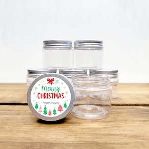 May include: A set of clear glass jars with silver lids. The jars are decorated with a round sticker that says "Merry Christmas" with a red bow and green Christmas trees. The sticker also says "From, Kevin".