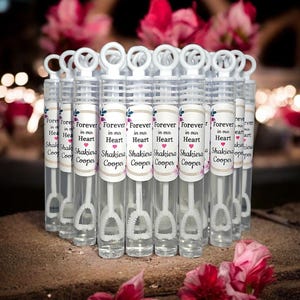 May include: A group of clear plastic bubble wands with white handles and labels that say "Forever in our Heart" with the names "Shakiera Cooper". The wands are arranged in a row on a brown surface with pink flowers in the foreground and background.