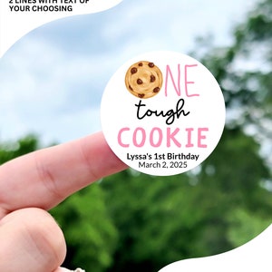 May include: A round sticker with a pink and white design. It features a cartoon cookie with chocolate chips and the text "ONE tough COOKIE". The sticker also includes the text "Lyssa's 1st Birthday March 2, 2025".