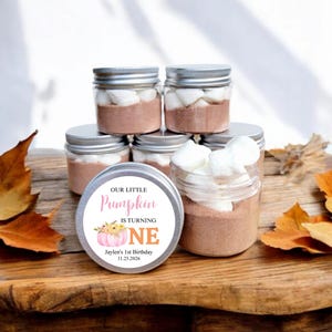 May include: A set of six small glass jars with silver lids, filled with hot chocolate mix and marshmallows. Each jar has a round sticker with a pink pumpkin and the text "Our Little Pumpkin is Turning One, Jaylen's 1st Birthday 11.25.2026".