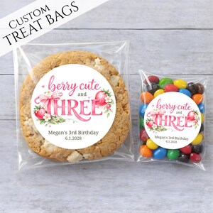 May include: Two clear treat bags, one with a large cookie and the other with colorful candies. Both bags have a circular sticker with the text "berry cute and THREE" and "Megan's 3rd Birthday 6.1.2028". The words "CUSTOM TREAT BAGS" are at the top.