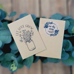 May include: A white card with the text "Let Love Grow" and a drawing of a mason jar filled with flowers. A second white card with a blue and white sticker that says "One day away" with the names Jack & Emily and the date 1.8.2023.