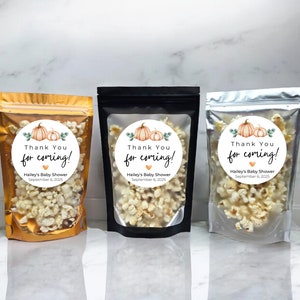 May include: Three clear plastic bags filled with popcorn, each with a round sticker featuring a watercolor pumpkin design and the text "Thank You for coming! Hailey's Baby Shower September 6, 2025". The bags are in gold, black, and silver.