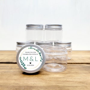 May include: A set of clear glass jars with silver lids. The jars are stacked on a wooden surface. One jar has a round sticker with the text "Thank you for celebrating with us! M&L 10.24.2024" in a gold circle with green leaves.