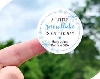 May include: A blue and white circle sticker with snowflakes and the text "A Little Snowflake Is On The Way" with a heart symbol. The sticker also includes the text "Baby Jones December 2026". The sticker is available in sizes 1 inch, 1.5 inches, 1.75 inches, 2 inches, 2.25 inches, and 3.33 inches. The sticker pictured is 2 inches.