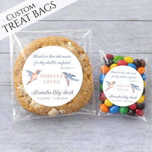 May include: Two clear treat bags, one with a large cookie and the other with colorful candies. Both bags have a circular label with text and hummingbird illustrations. The text reads "Blessed are those who mourn, for they shall be comforted, Forever Loved, Alexandra Riley Smith".