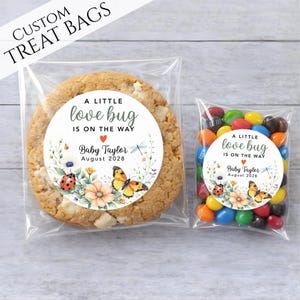 May include: Two clear plastic treat bags. One contains a large cookie with a white chocolate chip. The other contains colorful candies. Both bags have a circular sticker that reads "A little love bug is on the way" with a baby's name and date.