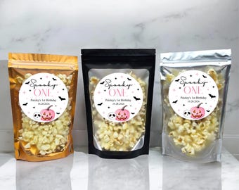 Op de afbeelding: Drie zakken popcorn met labels waarop "Spooky One" staat met een roze en zwarte pompoen en vleermuizen. De tekst op de labels zegt ook "Paisley's 1st Birthday" en een datum.