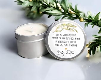 Op de afbeelding: Een zilveren kaarsenblik met een witte kaars en een deksel met tekst en een gouden bladeren ontwerp. De tekst luidt: "THIS IS A GIFT FOR YOU TODAY SO WHEN I'M BORN YOU'LL LIGHT MY WAY WITH THE FLICKER OF THE FLAME PLEASE SAY A PRAYER IN MY NAME Baby Furton."