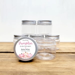 May include: A set of six clear glass jars with silver lids. The jars are labeled with a pink and white sticker that says "A Little Pumpkin is on the Way! Baby Miller February 2024". The sticker features a watercolor illustration of pink pumpkins.