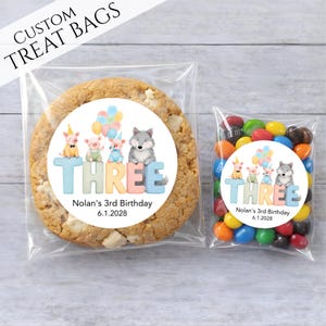 May include: Clear cellophane treat bags, one with a large cookie and the other with colorful candies. Both bags have a round sticker with a watercolor illustration of pigs, a wolf, balloons, and the word "THREE". Text reads "Nolan's 3rd Birthday 6.1.2028".