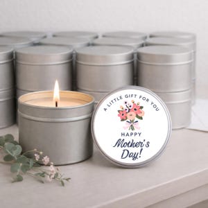 May include: A lit silver candle in a metal tin with a floral Mother's Day label. The label reads "A Little Gift For You" and "Happy Mother's Day!". Several similar candles are visible in the background.