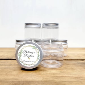 May include: Clear plastic jars with silver lids are stacked on a wooden surface. One jar has a decorative label with the text "Bethany's Baptism" and the date December 18, 2023, surrounded by a gold geometric design and green leaves.