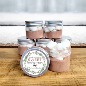 May include: Six clear glass jars with silver lids filled with hot chocolate mix and marshmallows. The jars have a white label with green and red holiday-themed graphics and the text "Wishing you a sweet holiday season. The Smith Family."