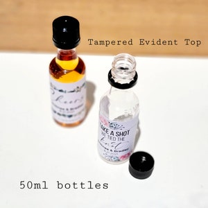 May include: Two 50ml clear glass bottles with black lids. One bottle is filled with a light amber liquid and has a white label with the text "Cheers" and a floral design. The other bottle is empty and has a white label with the text "Take a shot we tied the knot" and a floral design. The text "Cathee & Bradley 8.21.2021" is also on the label.
