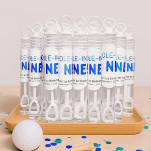 May include: A collection of clear bubble wands with white handles and blue text that reads "NINE". The wands are arranged on a wooden tray, with a golf ball, tee, and confetti nearby. A banner of blue and green triangles is in the background.