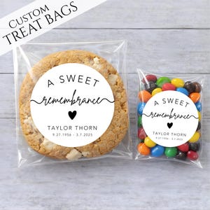 May include: Two clear plastic treat bags, one with a large cookie and the other with colorful candies. Both bags have a white circular label with the text "A SWEET remembrance", a heart, and a name with dates. The words "CUSTOM TREAT BAGS" are at the top.