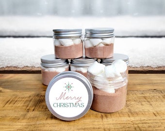 May include: Six clear glass jars filled with hot chocolate mix and marshmallows. The jars have silver lids and are stacked in a pyramid shape. The jar in the front has a white label with the text "Merry Christmas" and a green snowflake design.